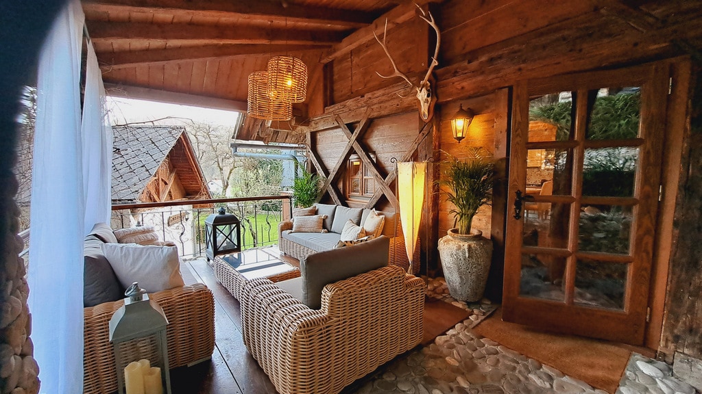 A relaxing outdoor seating area is presented with comfortable wicker furniture under a wooden roof. Soft lighting is provided by wall sconces and a decorative lantern. Greenery adds a natural touch, while stone flooring complements the rustic aesthetic.