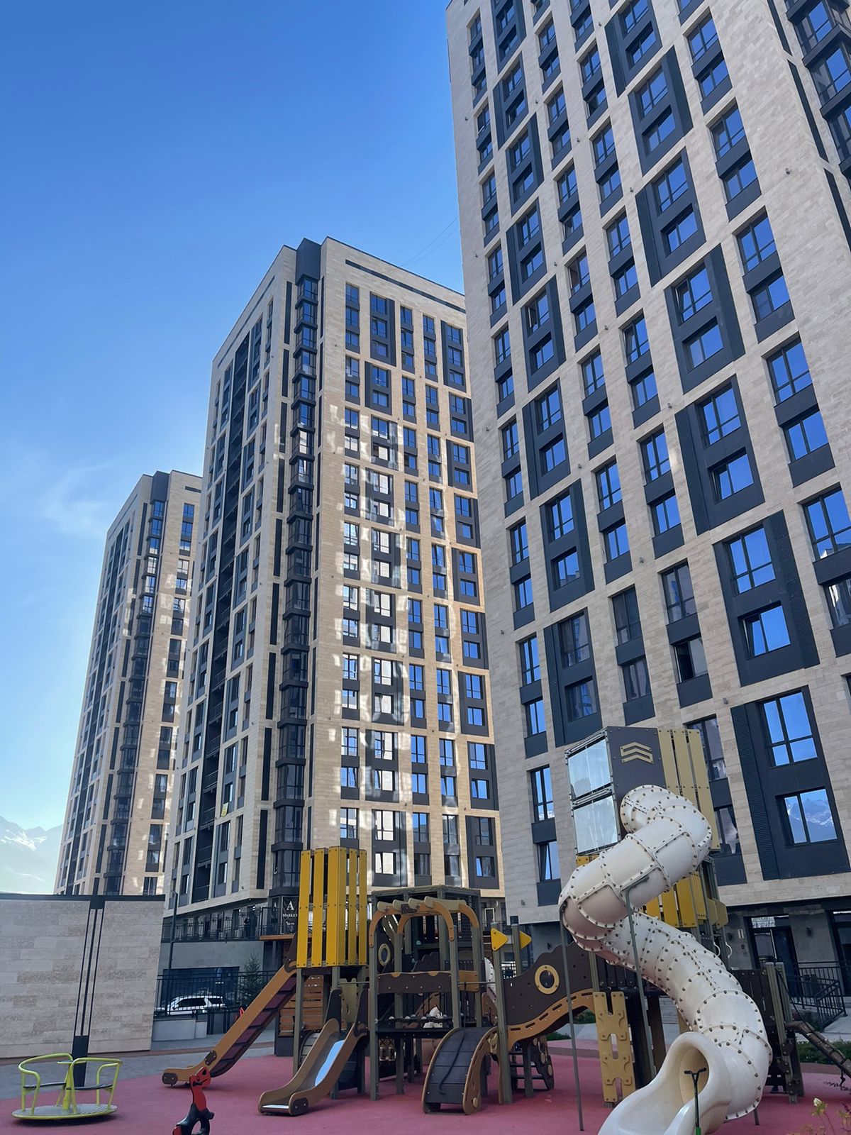 Modern residential buildings are showcased, characterized by large, reflective windows. The foreground features a playground with a slide and climbing structure, situated on bright red surfacing. Clear blue skies create a welcoming atmosphere around the buildings.