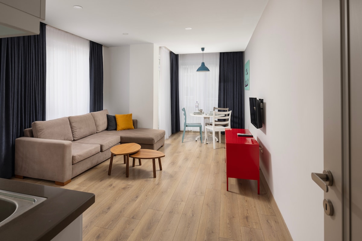 A well-lit living space features a comfortable sofa paired with an accent pillow. A circular wooden coffee table is positioned in front of the sofa. A dining area with a white table and chairs is visible, along with a vibrant red storage unit and a wall-mounted television.