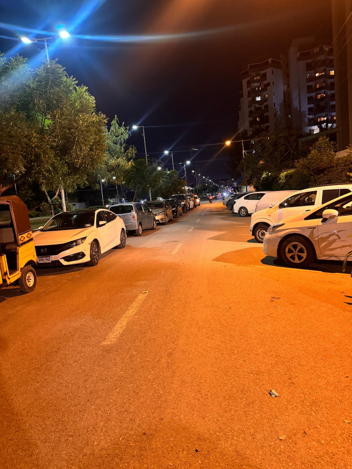 A well-lit street is lined with parked cars on both sides, showcasing a variety of vehicles under bright streetlights. Tall trees are visible along the street, with apartment buildings illuminated in the background.