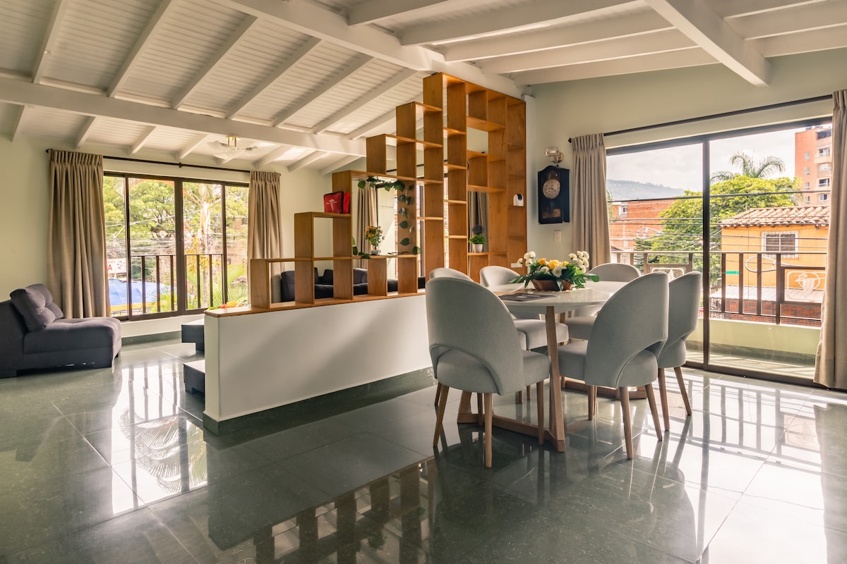 A spacious dining area features a round table surrounded by upholstered chairs. Large windows provide natural light and views of the outdoors, while a stylish wooden shelf adds warmth and structure to the room. The green tile floor reflects the inviting atmosphere.
