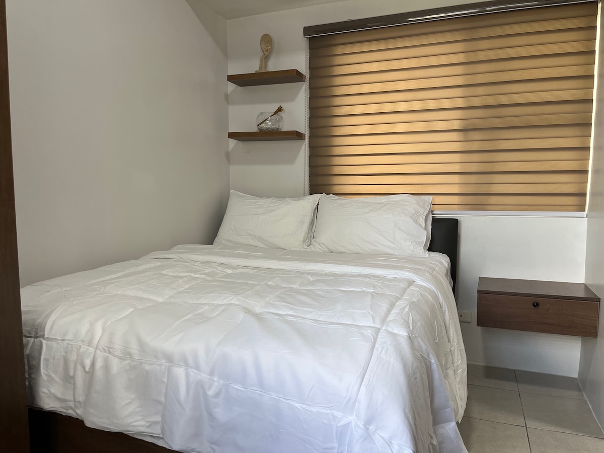 A spacious sleeping area features a large bed topped with white linens, set against a neutral wall. Natural light filters through a wooden blinds arrangement, and a small nightstand with a decorative item can be seen nearby.