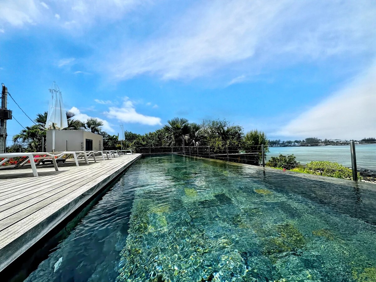 An infinity pool stretches towards the horizon, surrounded by a wooden deck equipped with lounge chairs. Lush greenery and coastal views are seen in the background, under a clear blue sky with light clouds.