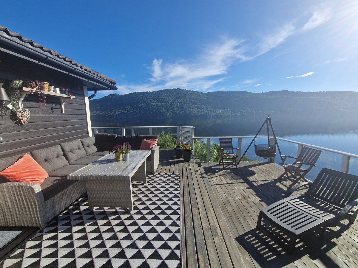 A spacious terrace features comfortable seating arrangements, including a lounge sofa and deck chairs. Scenic views of the fjord and surrounding mountains are visible, framed by clear skies. Decorative plants and a patterned rug enhance the outdoor space, inviting relaxation and enjoyment of the natural beauty.