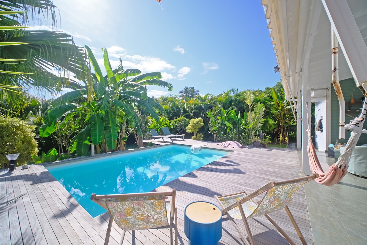 A refreshing pool is framed by lush greenery and palm trees, creating a tropical ambiance. Wooden decking surrounds the pool, featuring two chairs and a small table. A hammock is visible, inviting relaxation in a serene outdoor space under clear blue skies.