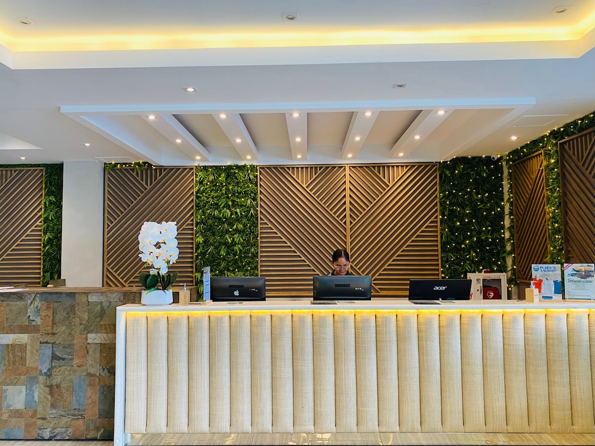 The reception area features a sleek front desk illuminated by soft lighting. A green wall adorned with plants adds a refreshing touch, while the stone and wood elements provide a modern and inviting atmosphere. Informational displays are neatly arranged on the countertop.