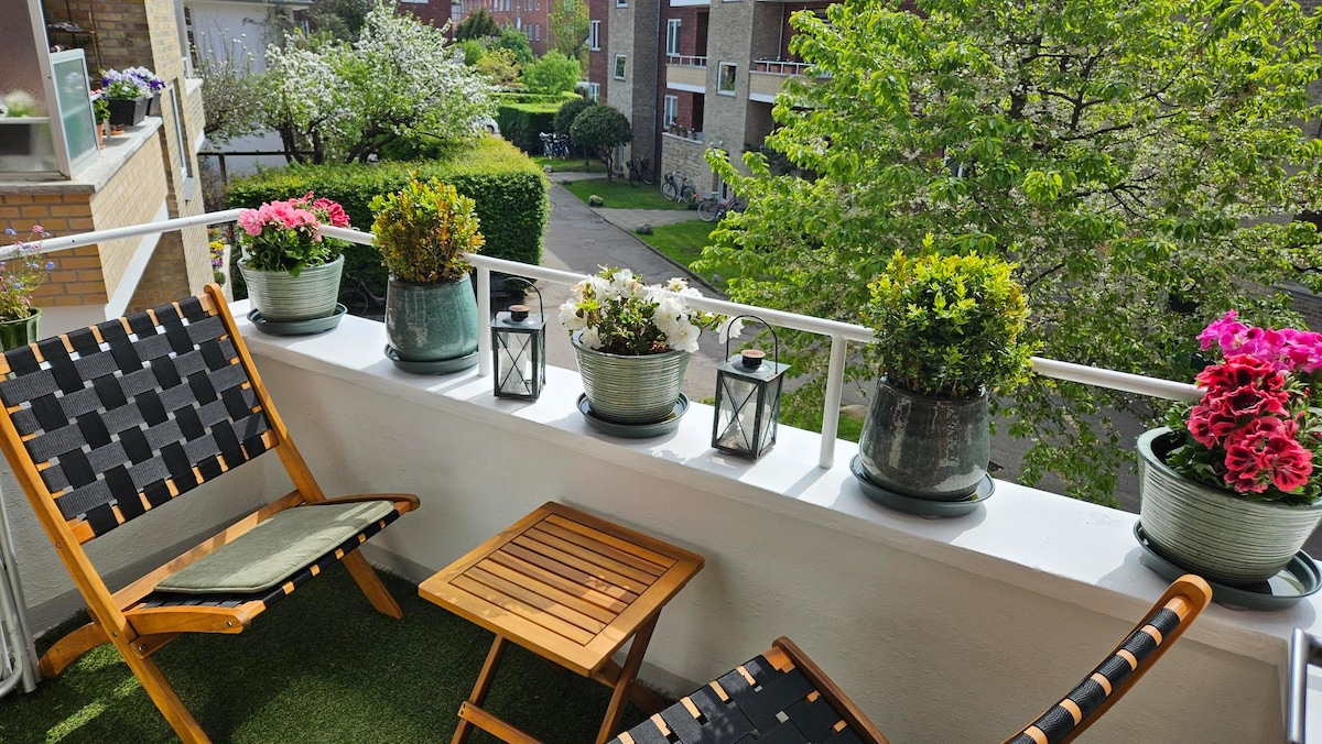 A balcony is presented with two wooden chairs positioned beside a small, foldable table. Several planters filled with vibrant flowers and greenery line the railing, creating a peaceful outdoor space. The view includes lush trees and landscaped surroundings.