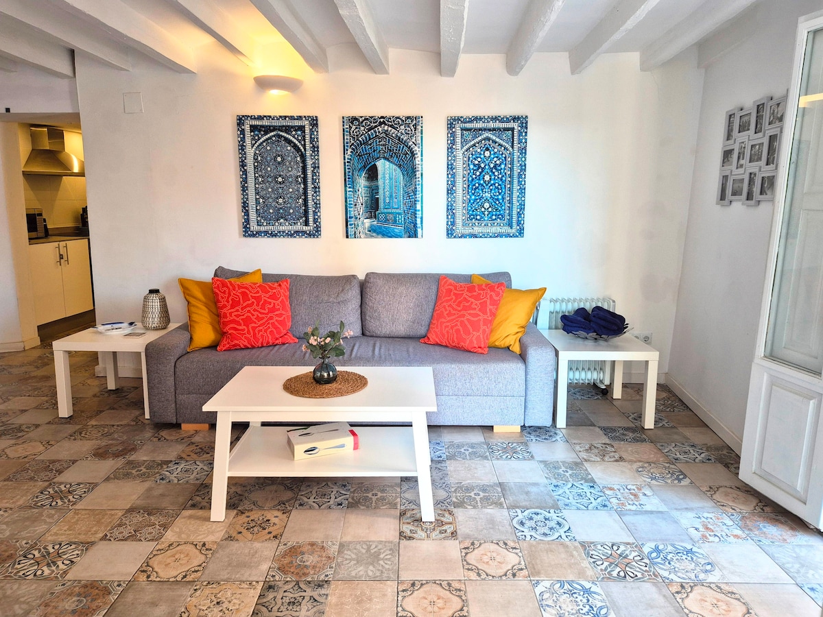 A cozy living area features a gray sofa adorned with vibrant orange and yellow cushions. A white coffee table sits on patterned tile flooring, complemented by decorative wall art showcasing intricate blue designs. A glimpse of the kitchen area is visible in the background.