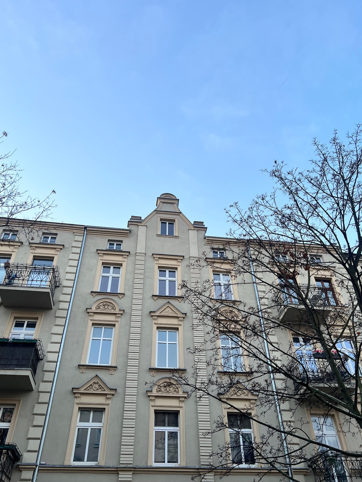 The historic building exterior is displayed against a clear blue sky. Architectural details, including decorative moldings and multiple windows, are visible. Trees frame the scene, adding natural elements to the urban landscape.