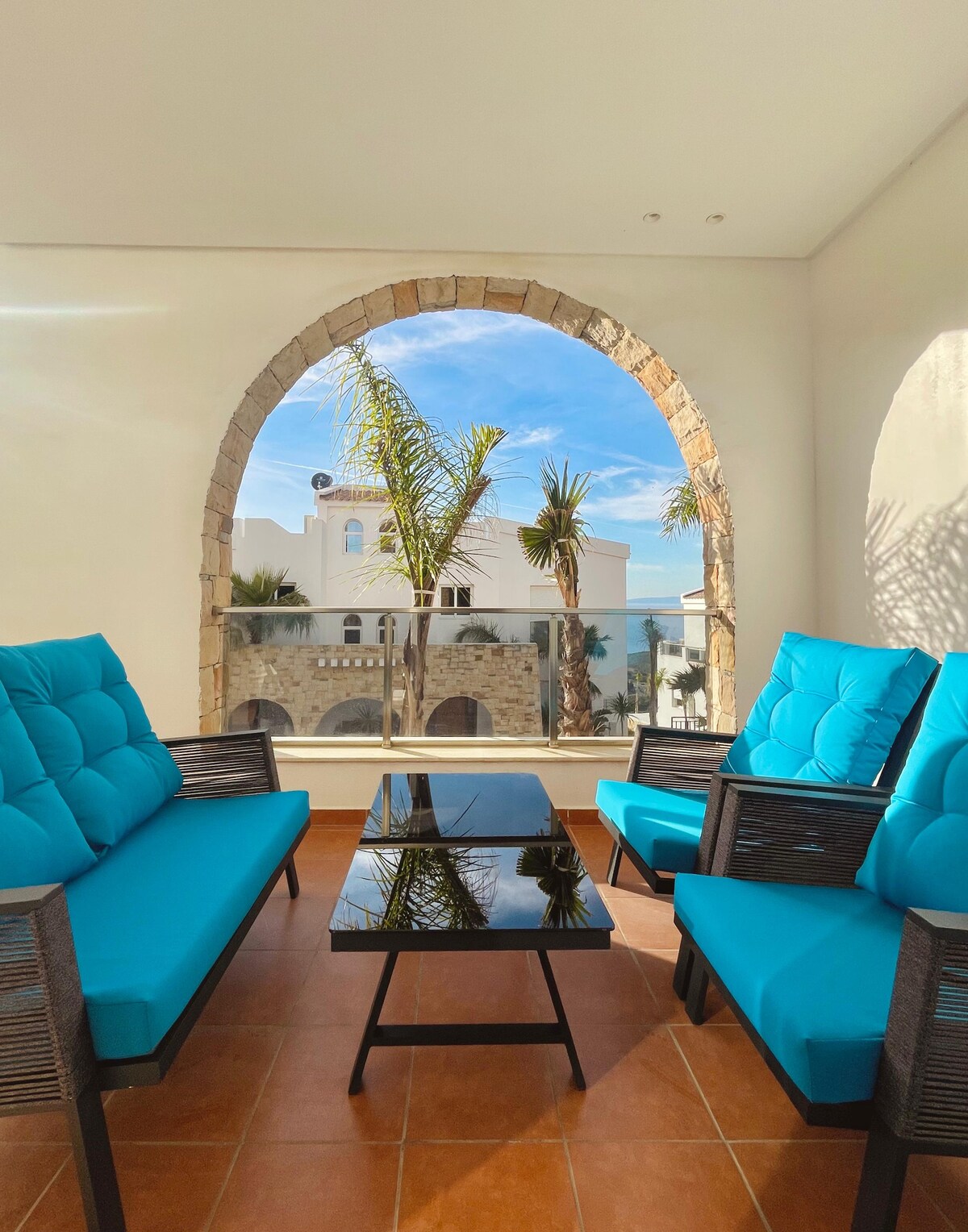 An arched stone window frames a view of palm trees and a distant building. Colorful blue seating arranged around a sleek black table creates a comfortable gathering area. Natural light enhances the spacious atmosphere, reflecting softly off the tiled floor.