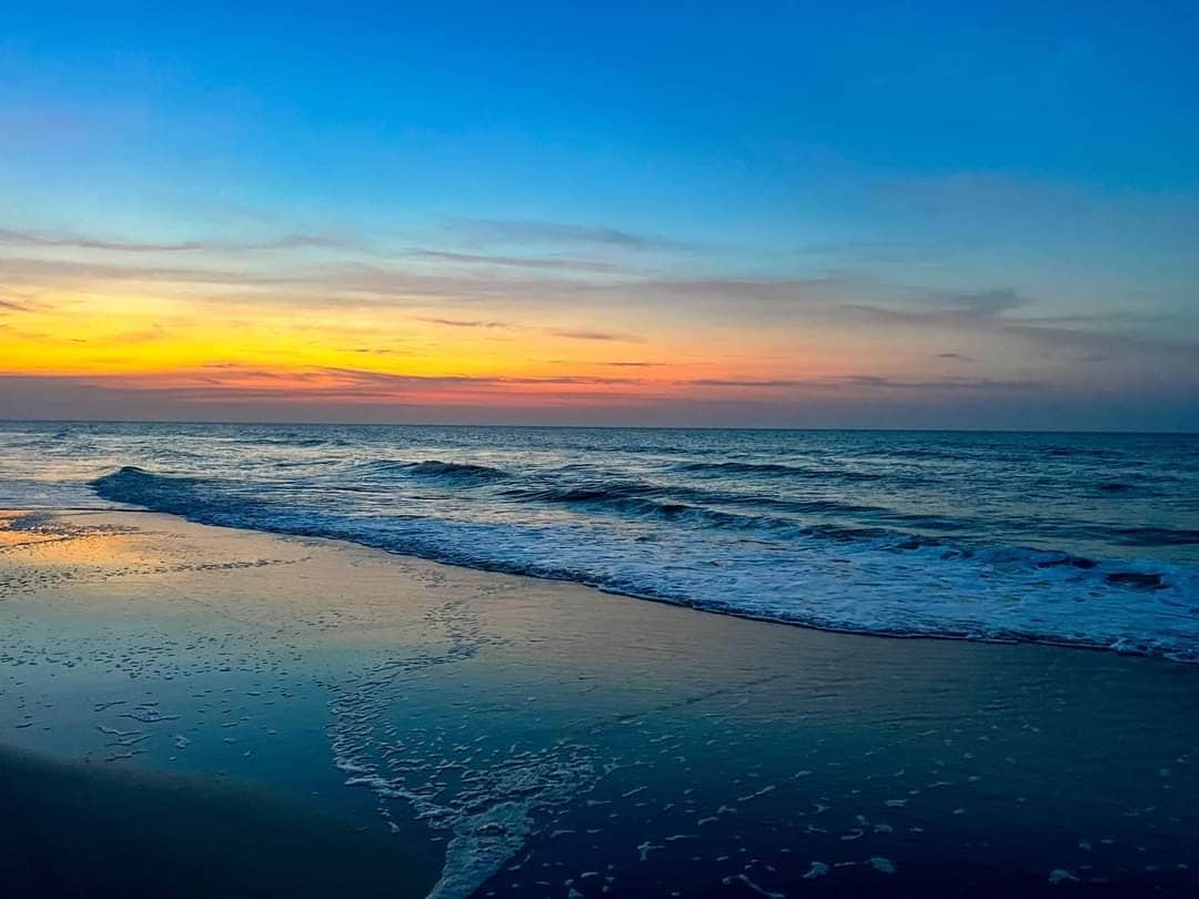 The image captures a serene beach scene at twilight, where gentle waves meet the shore. The sky displays a gradient of warm colors transitioning from orange to deep blue, reflecting softly on the wet sand.