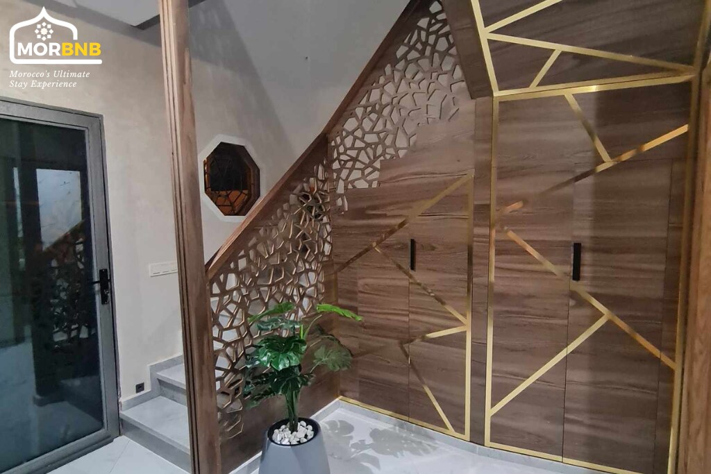 An intricately designed staircase features wooden paneling with geometric cutouts, complemented by elegant gold accents. A potted plant adds a touch of greenery, and the adjacent wall contains a decorative storage unit. Natural light filters in through a nearby glass door.