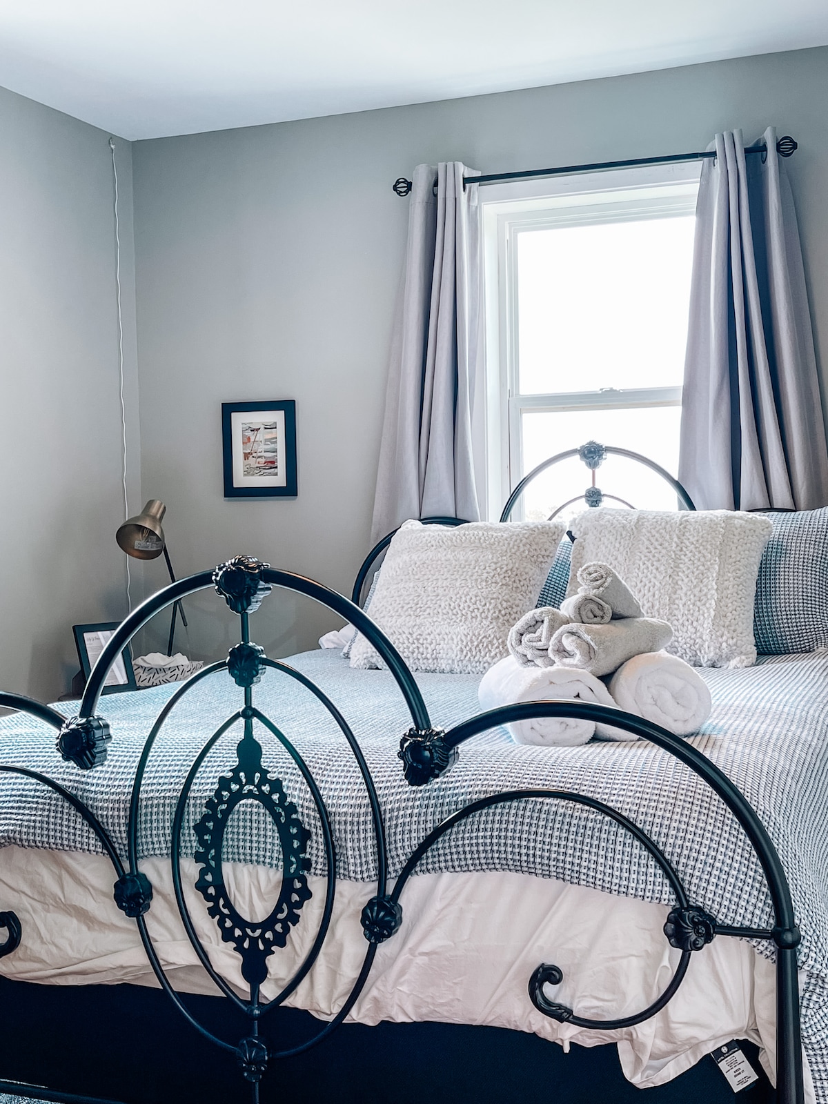 A comfortable bedroom features a wrought-iron bed frame adorned with decorative pillows and neatly rolled towels. Light filters through a window, surrounded by coordinating drapes. A bedside lamp and a framed picture add to the inviting ambiance of the space.