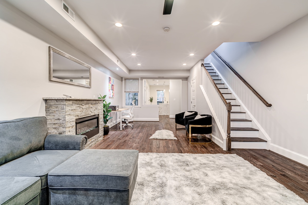 The spacious living area features a plush gray sectional sofa near a stone-accented fireplace. A soft area rug lies on the hardwood floor, complementing the minimalist decor. A desk is positioned near a large window, with a staircase leading to the upper level in the background.