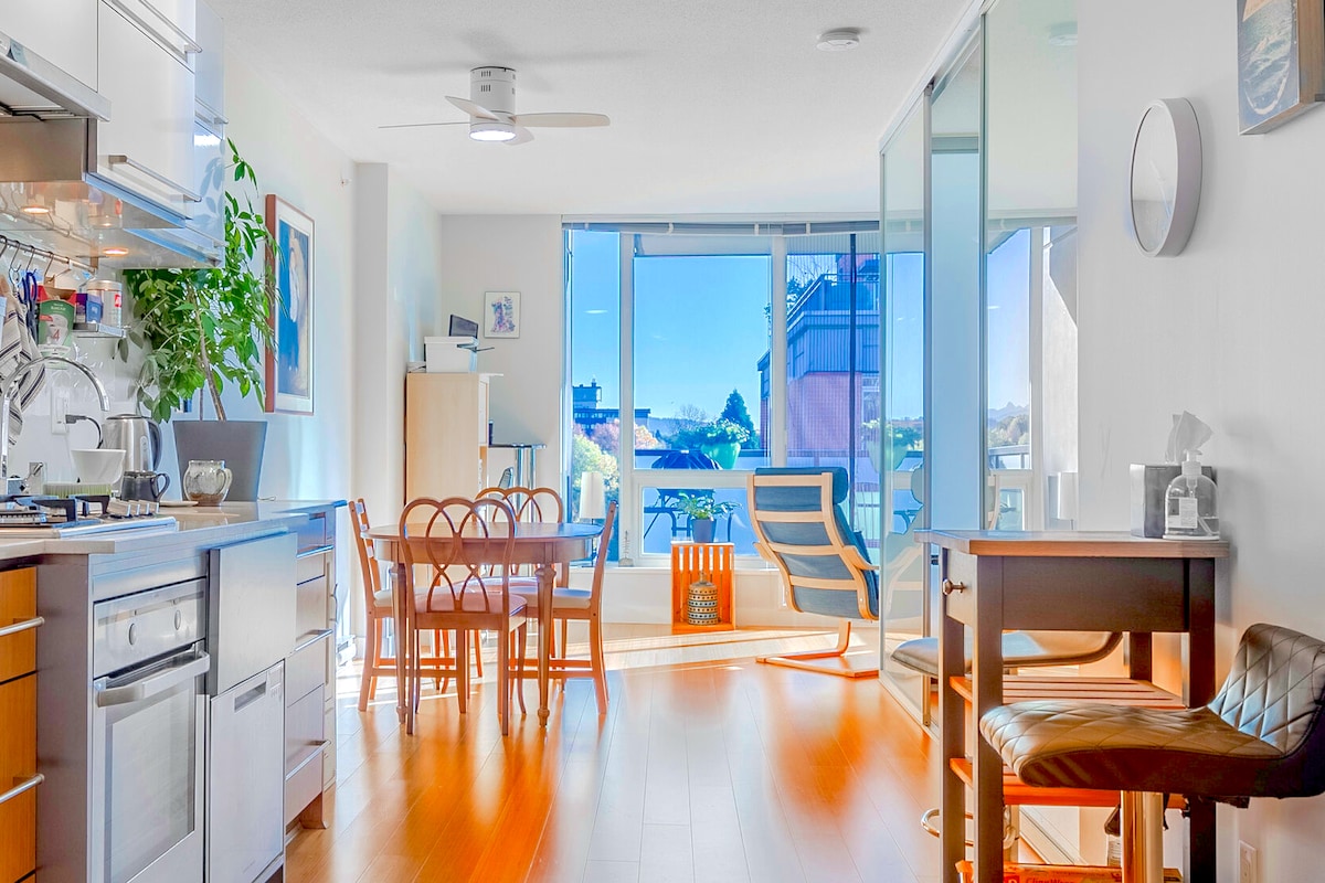 A bright and airy living space features large windows allowing plenty of natural light. The kitchen, dining area, and workspace are well-defined, with a table for four, a standing desk, and comfortable seating. The balcony is visible in the background, enhancing the overall openness.