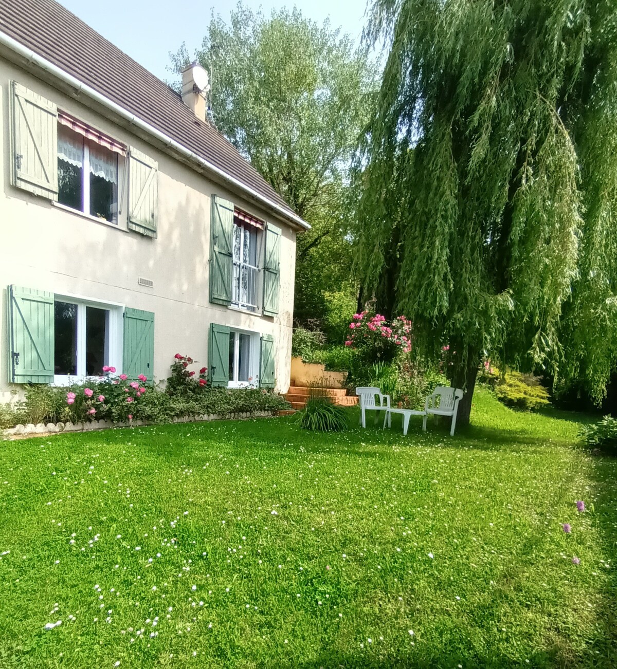 A well-maintained exterior of a home is depicted, featuring green shutters and white walls surrounded by a lush green lawn. Two white chairs are positioned on the grass near blooming flowers, with trees providing shade and contributing to a serene outdoor environment.