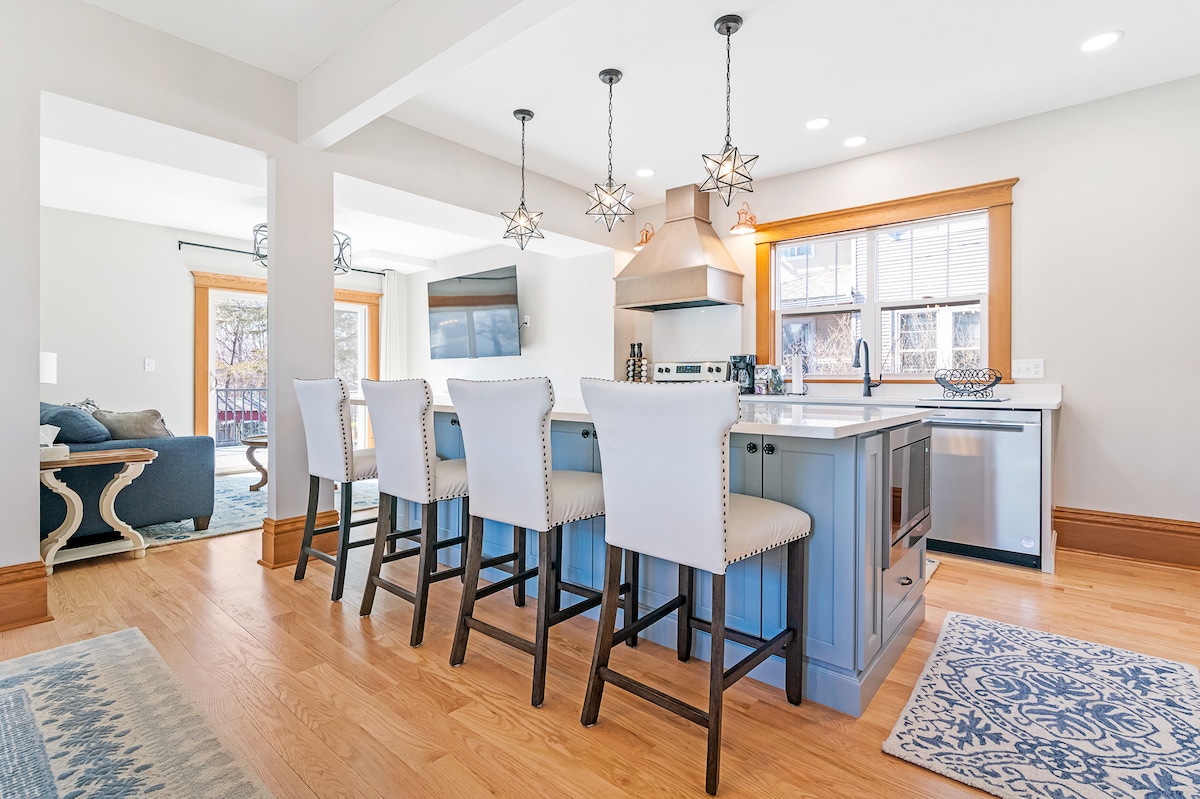 A spacious kitchen area features a central island with seating for six underneath decorative star-shaped pendant lights. Stainless steel appliances and a bar refrigerator are visible. Natural light floods the room through multiple windows, illuminating the hardwood floor and neutral-colored cabinetry.