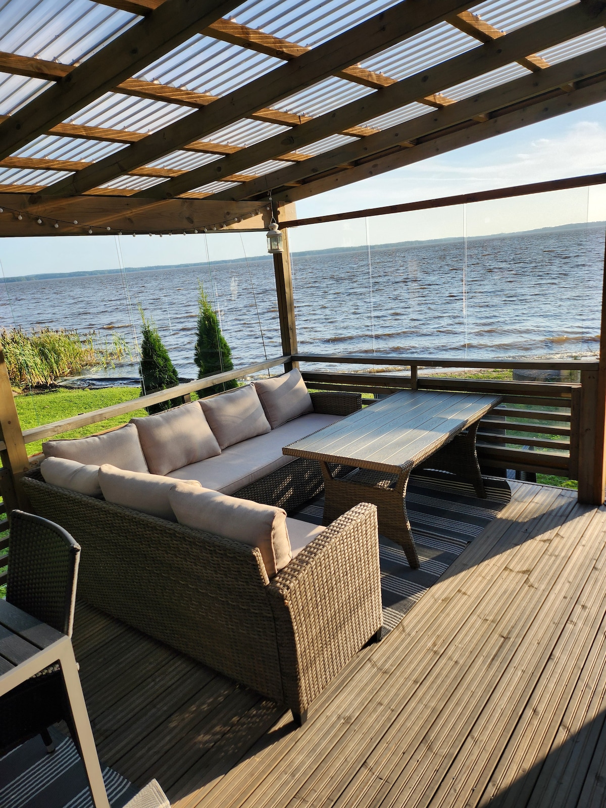 A shaded terrace overlooks the tranquil waters, featuring comfortable wicker seating with plush cushions. A wooden table sits between the seating area, providing a space for relaxation or dining. The scene is framed by greenery and the shimmering surface of the lake.