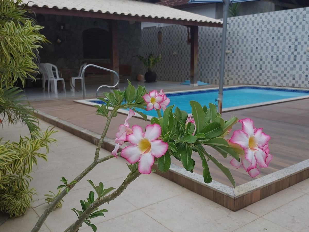 A vibrant flowering plant is positioned in the foreground, showcasing pink and white blossoms. In the background, a tiled pool reflects the surrounding patio, which is bordered by a shaded area with outdoor seating. The space offers a blend of natural beauty and leisure.