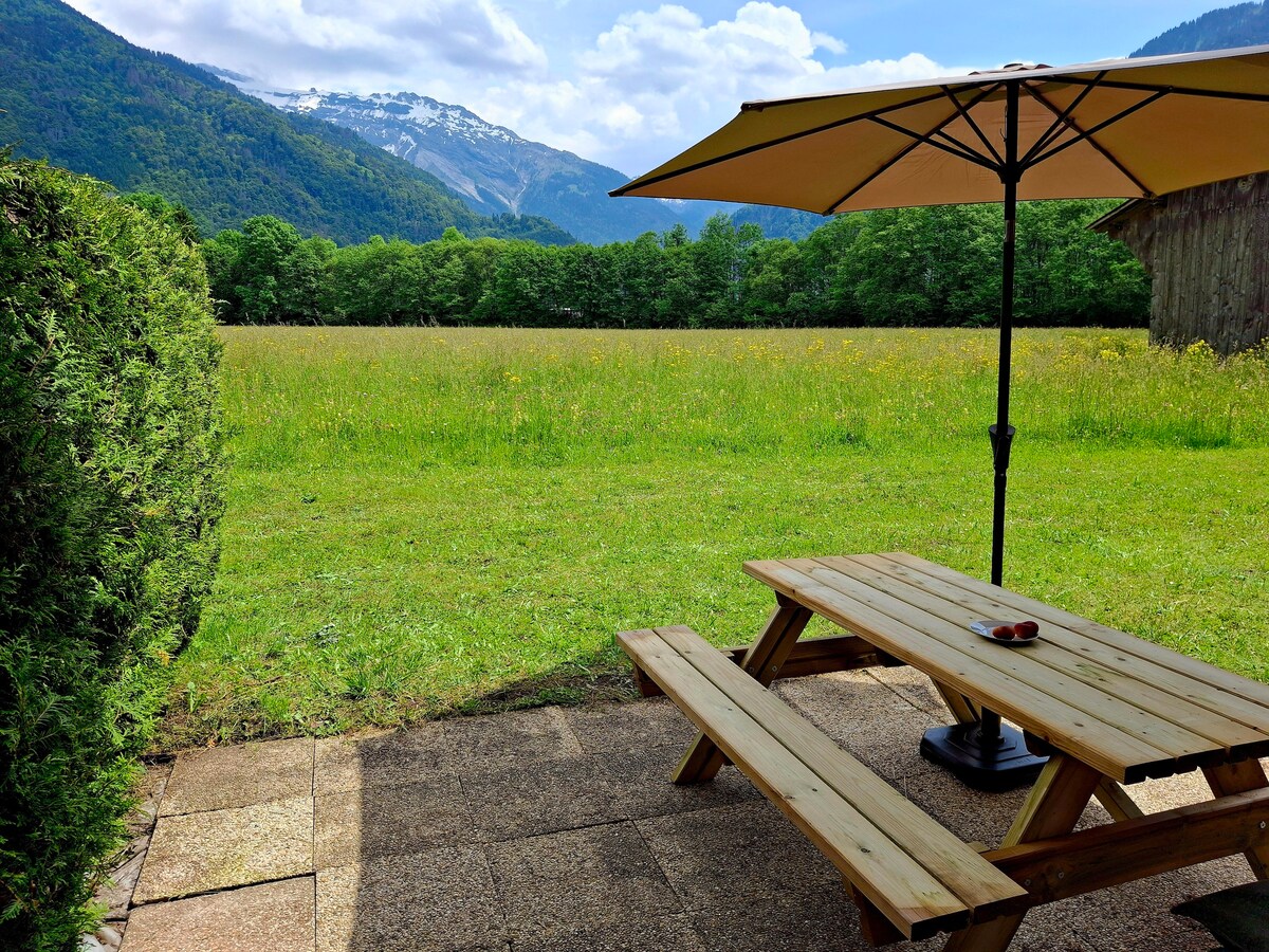 A spacious terrace features a picnic table with benches, shaded by a large umbrella. A grassy field extends beyond, framed by lush trees and mountains in the distance. The sunny atmosphere invites relaxation while enjoying the natural surroundings.