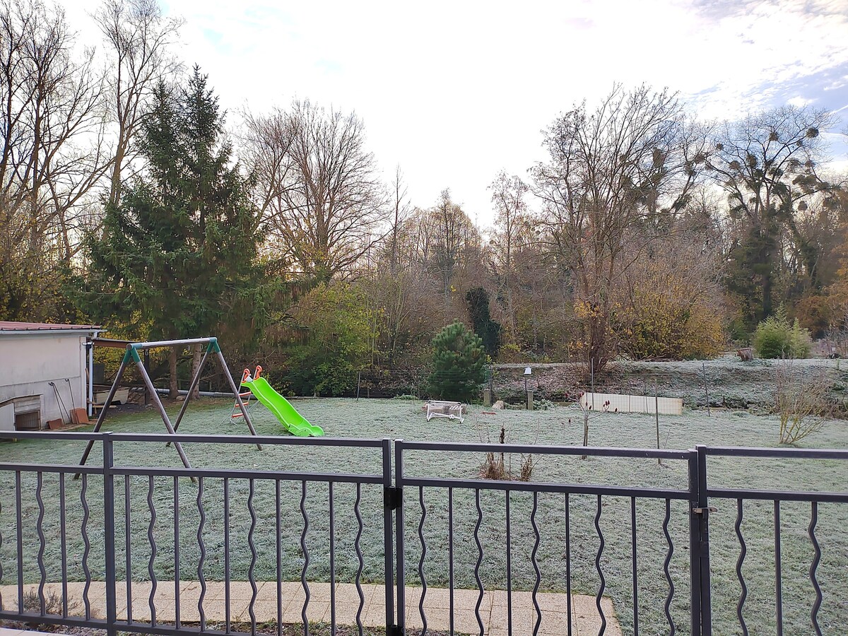 A spacious backyard is presented, featuring a swing set and a green slide. Frost covers the grass, surrounded by trees and shrubs in a serene environment. A distant wooden bench is visible, enhancing the outdoor setting.
