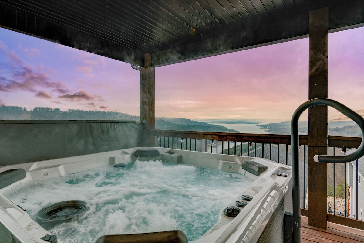A large hot tub is situated on the top deck, surrounded by wooden railings. Steam rises from the bubbling water, contrasting against the colorful sunset sky, showcasing hues of pink and purple over Table Rock Lake in the background.
