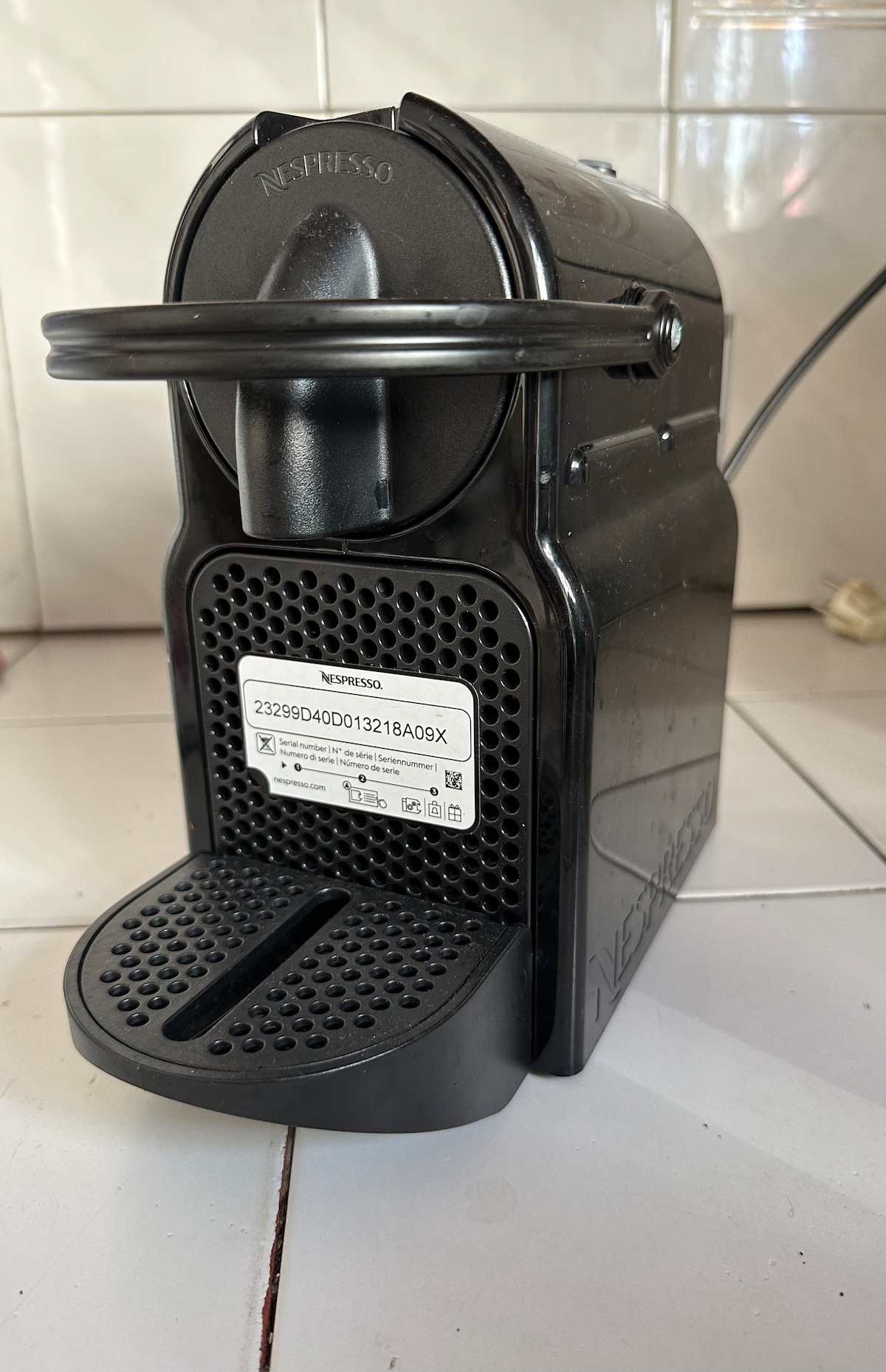 A modern coffee machine is positioned on a tiled countertop. The black body features a round handle and a grid surface for securing cups, with a small label visible on the front. The kitchen backdrop consists of white tiles.