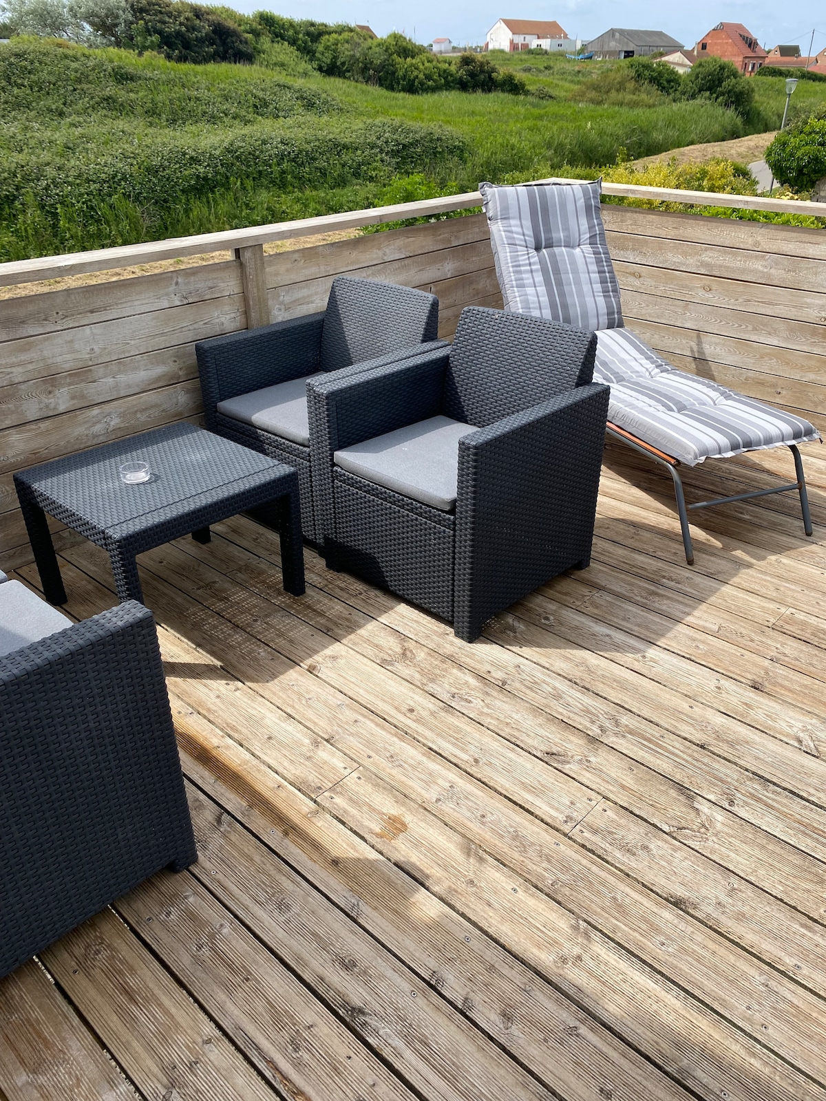 An outdoor terrace is furnished with two modern lounge chairs and a small table. The flooring is made of wooden planks, providing a natural setting. Lush greenery is visible in the background, enhancing the tranquil outdoor environment.