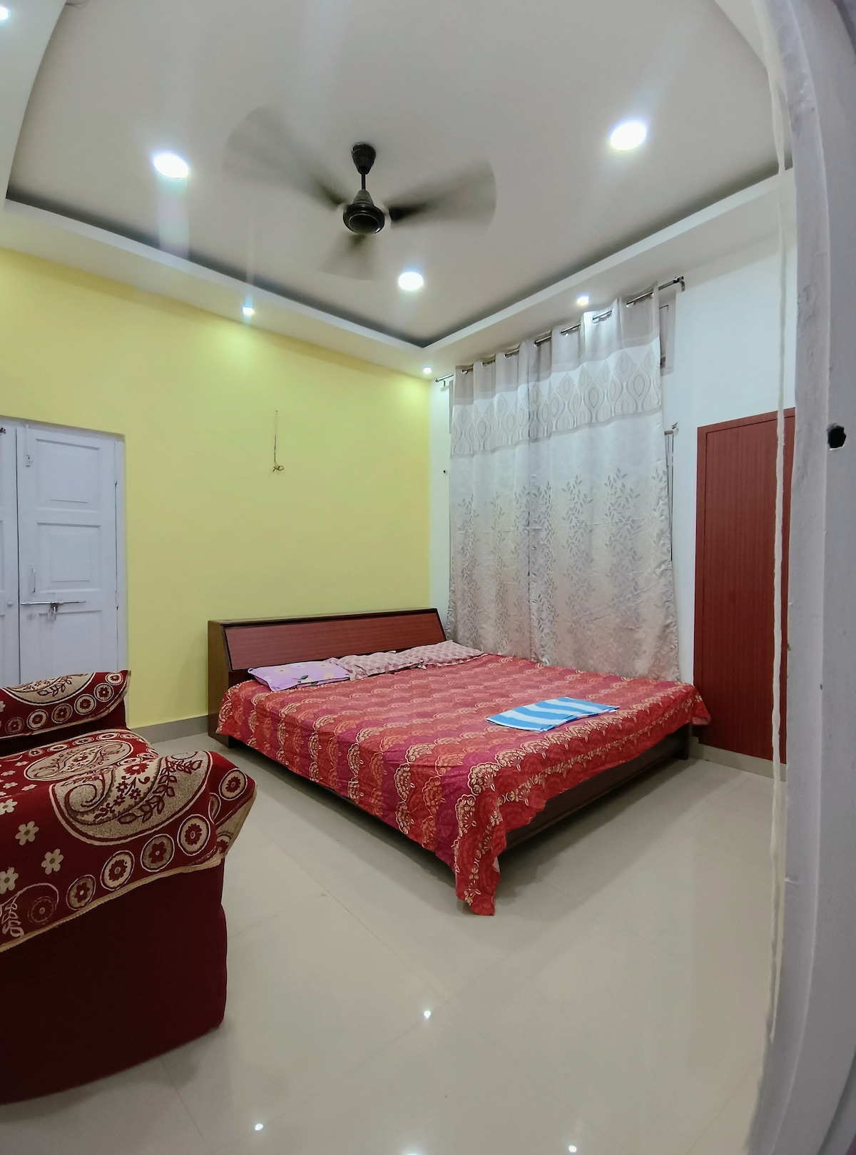 A well-lit bedroom features a spacious bed covered with a red patterned blanket. A cozy seating area is presented with a comfortable chair. Light curtains softly diffuse natural light, while the walls are painted a cheerful yellow, enhancing the room's overall brightness.