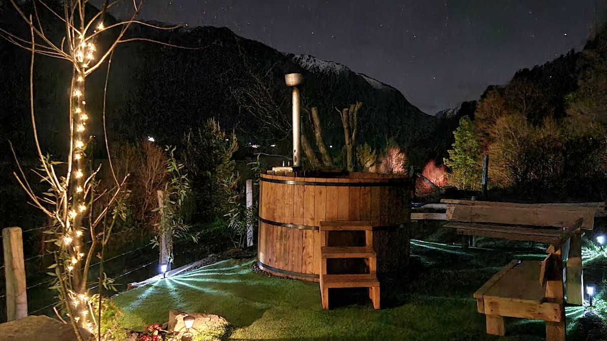 An outdoor area is illuminated by soft lights, showcasing a wooden hot tub surrounded by nature. A staircase leads up to the tub, and seating is available nearby. In the background, mountains are visible under a night sky, enhancing the tranquil atmosphere.