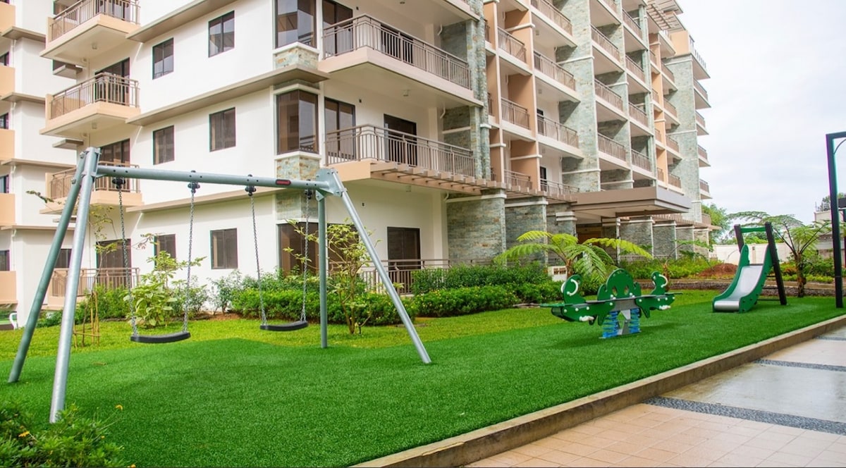 The exterior of the condominium complex is featured, showcasing a well-maintained green lawn with play equipment including a swing set and a multi-use play structure. Residents can enjoy a serene environment with greenery surrounding the area, complemented by the modern architectural design of the building.