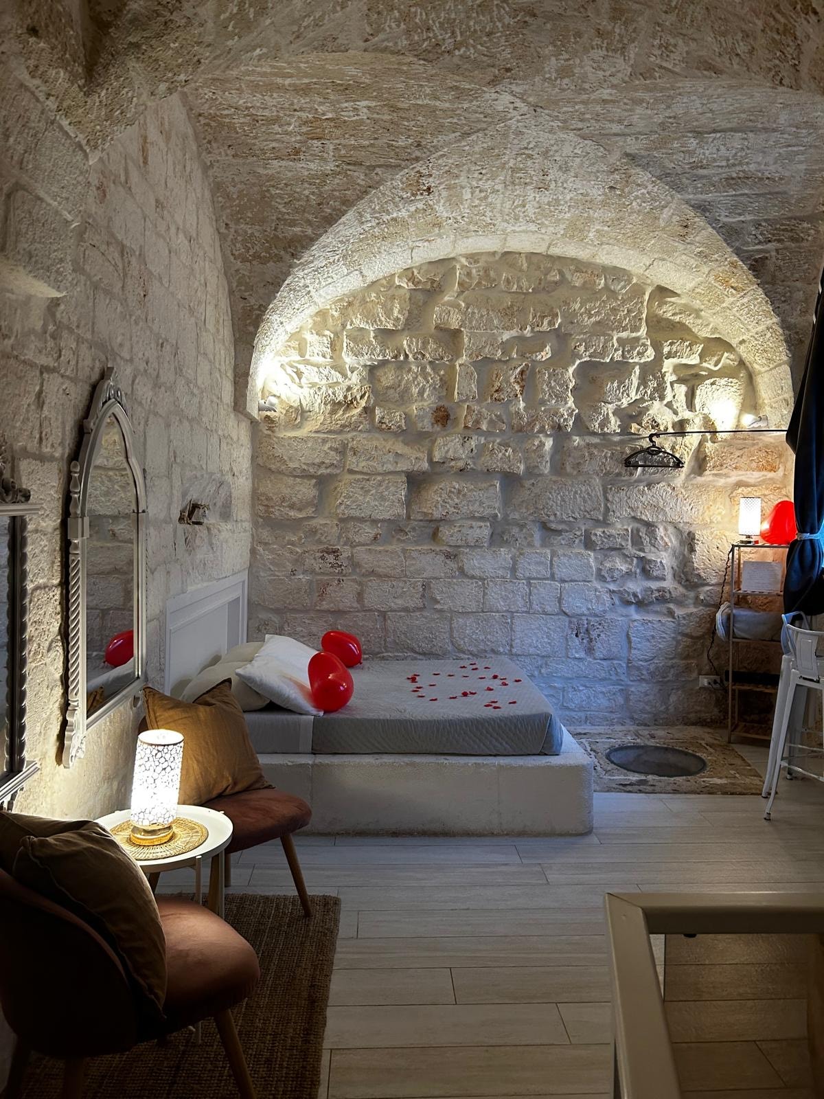 A cozy sleeping area features an arched stone ceiling and exposed stone walls. A simple bed is adorned with decorative red balloons and rose petals. Complementary furnishings include two chairs and a small table with a lamp, creating an intimate atmosphere.
