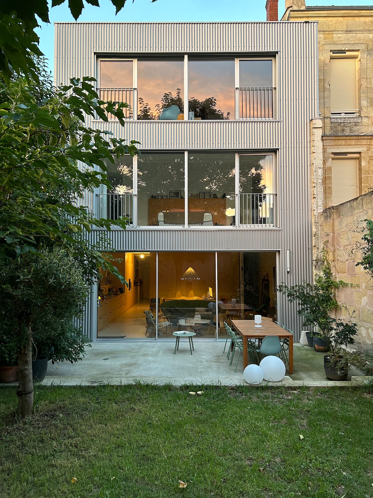 The rear view of the modern house reveals large glass panels reflecting the sky, providing access to the spacious living area and garden. A wooden dining table is set on the patio, surrounded by greenery. Soft garden lighting enhances the tranquil atmosphere.