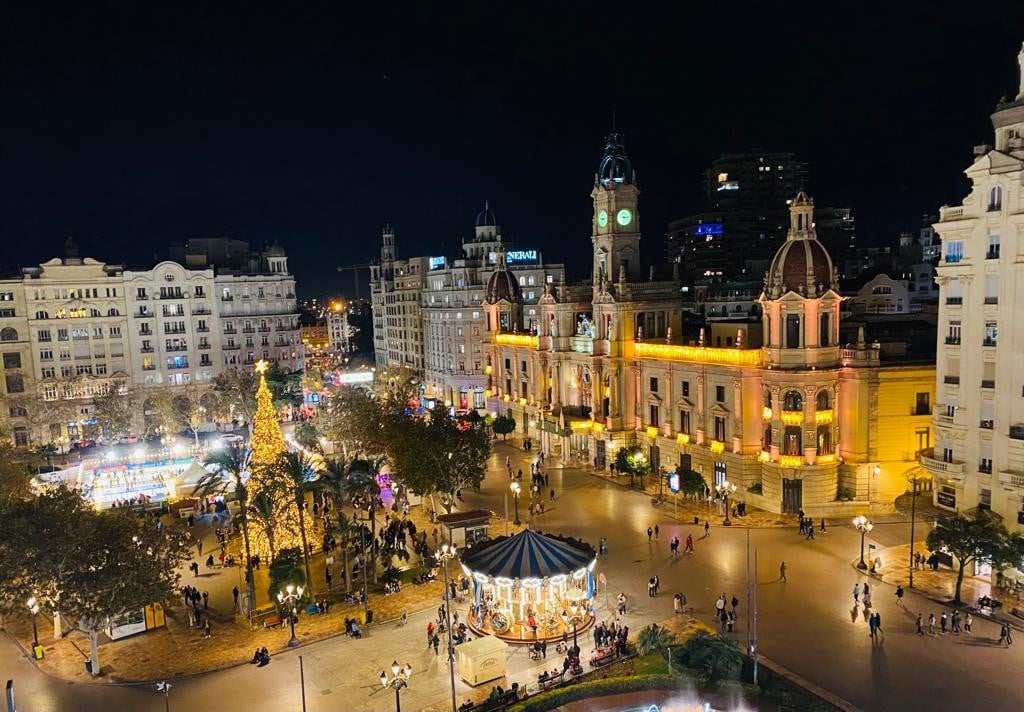 The image captures a lively square at night, illuminated by lights. A festive carousel is visible in the foreground, surrounded by palm trees. The historic buildings, including a clock tower, add an elegant backdrop, with colorful decorations enhancing the festive atmosphere.