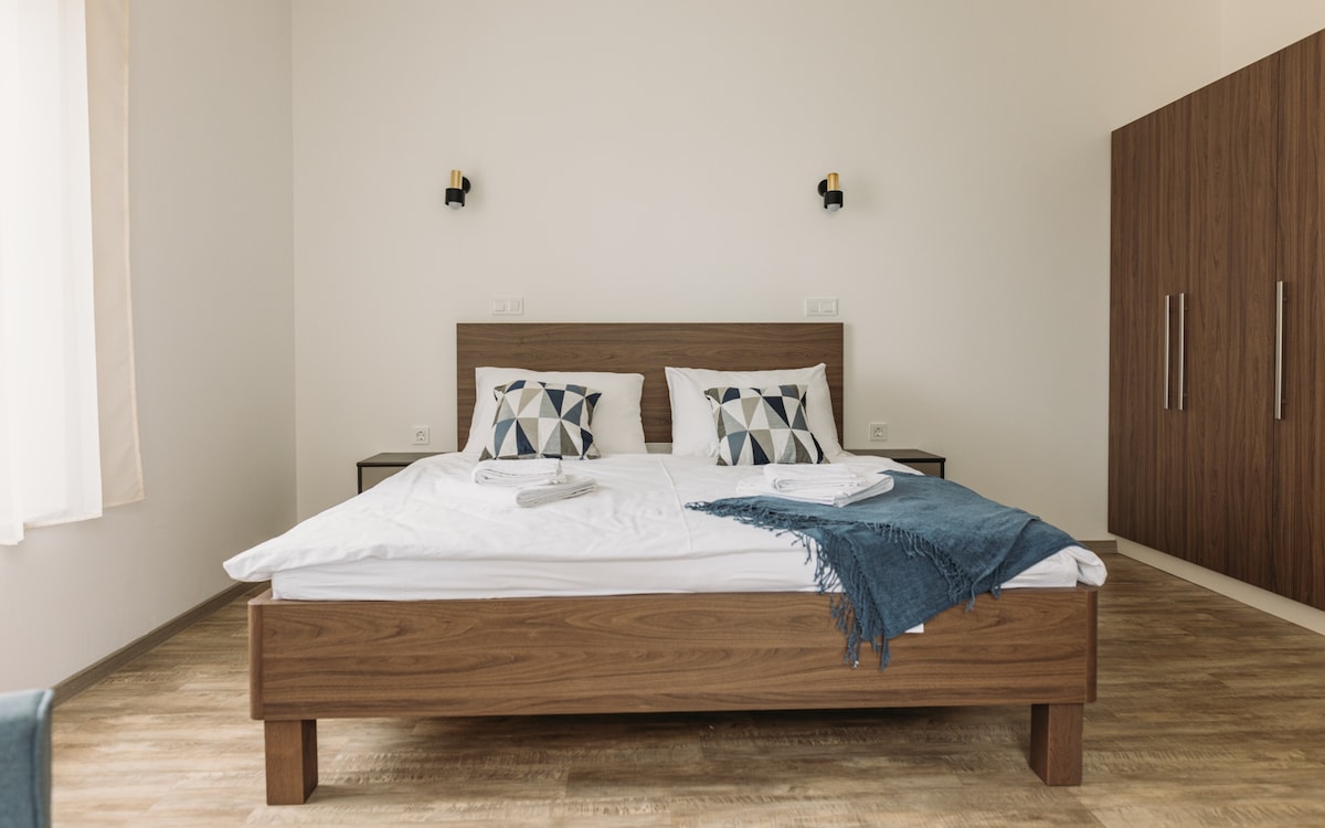 A spacious bedroom features a wooden bedframe with a neatly arranged white comforter and accent pillows adorned with geometric patterns. A cozy blue throw is draped over the bed. Complementary wooden furnishings include a wardrobe, enhancing the room’s clean, modern aesthetic.