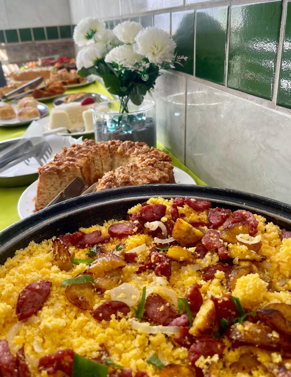 A large dish of traditional Brazilian food is displayed, featuring a vibrant mix of yellow grains topped with colorful vegetables and sliced sausages. In the background, a bouquet of white flowers sits on a table, alongside various pastries and a cake, enhancing the inviting dining atmosphere.