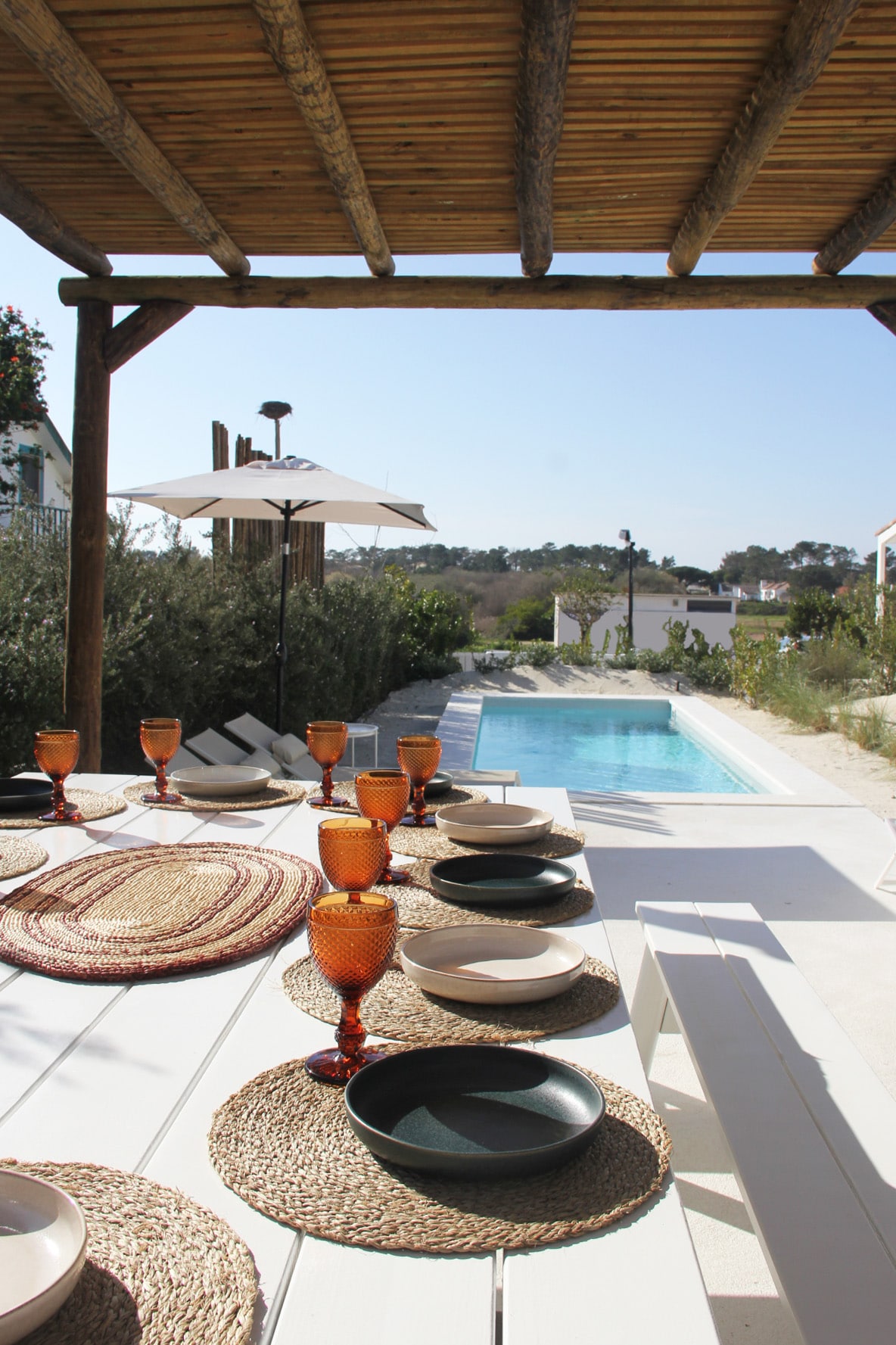 An outdoor dining area features a long white table set for a meal, adorned with colorful glasses and plates. Beyond the table, a private swimming pool glistens in the sunlight, surrounded by greenery and lounge chairs, with a sun umbrella providing shade.