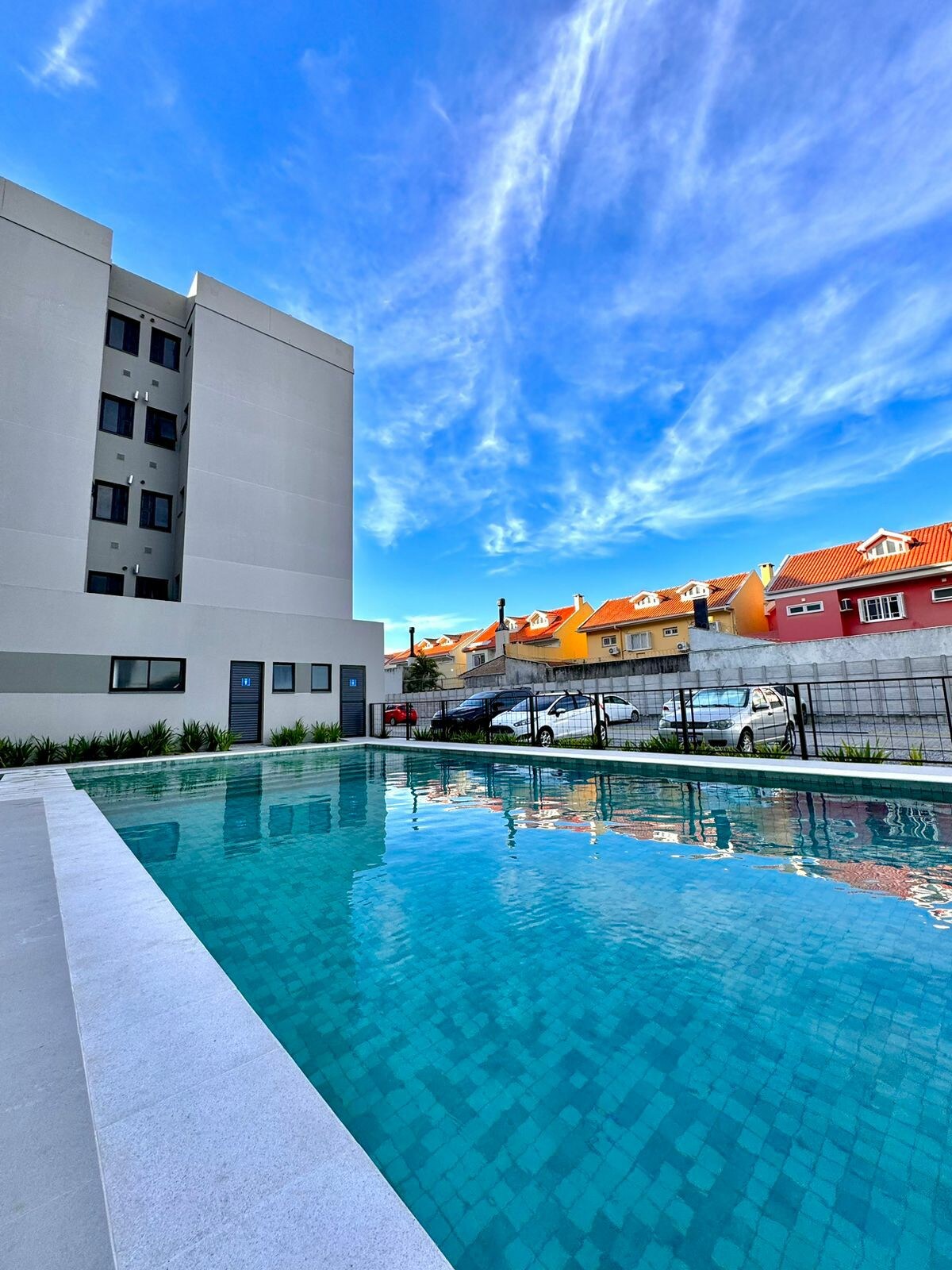 A serene swimming pool is surrounded by a clean, well-maintained area, reflecting a clear blue sky. The modern building stands nearby, with several vehicles visible in the background, showcasing the convenience of on-site parking.