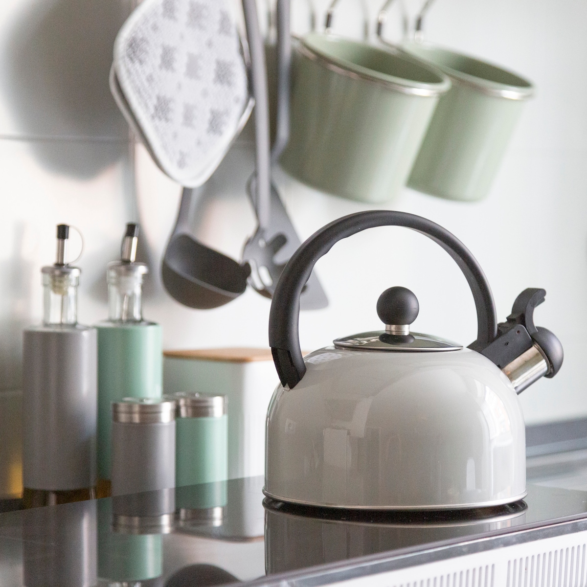A sleek kettle sits on a polished stovetop, reflecting natural light. Surrounding it are neatly arranged kitchen utensils, including a pair of mint-green containers, measuring spoons, and a grey pot holder, contributing to a well-organized cooking space.