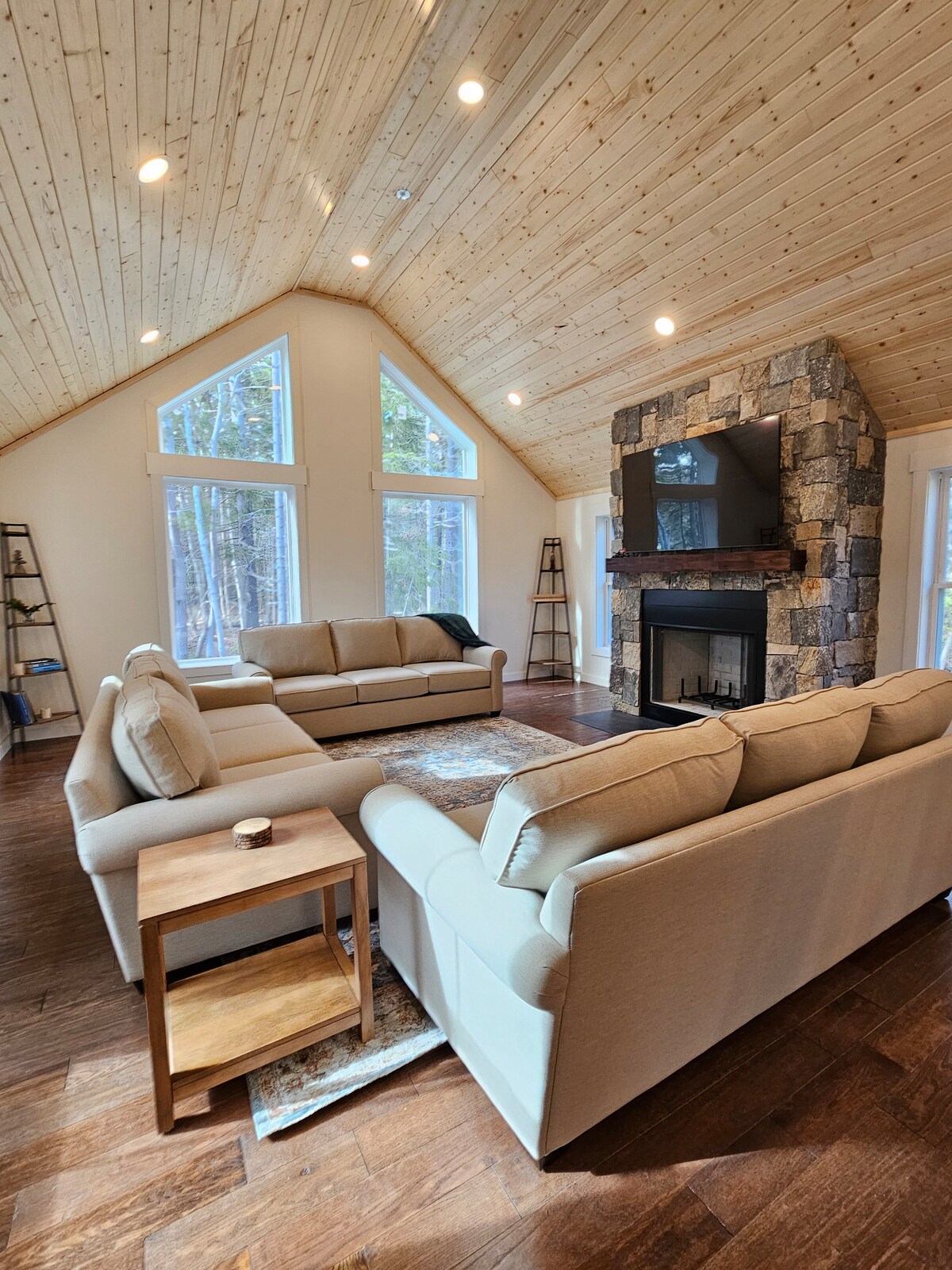 A spacious great room features a vaulted wood-paneled ceiling and large windows that provide views of the surrounding woods. Two beige sofas are arranged around a decorative area rug, with a stone fireplace and mounted television as a central focal point.