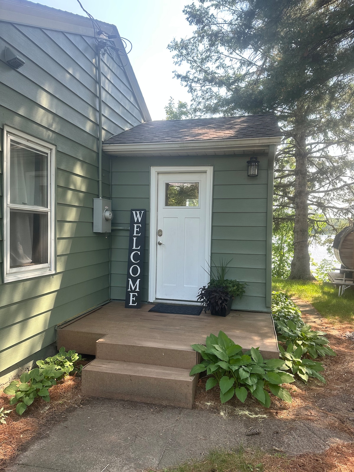 The entrance to the cabin is depicted, featuring a welcoming sign beside a white door. Steps lead up to the entrance, framed by green siding and landscaping, including leafy plants along the path. Trees provide shade in the background, enhancing the natural setting.