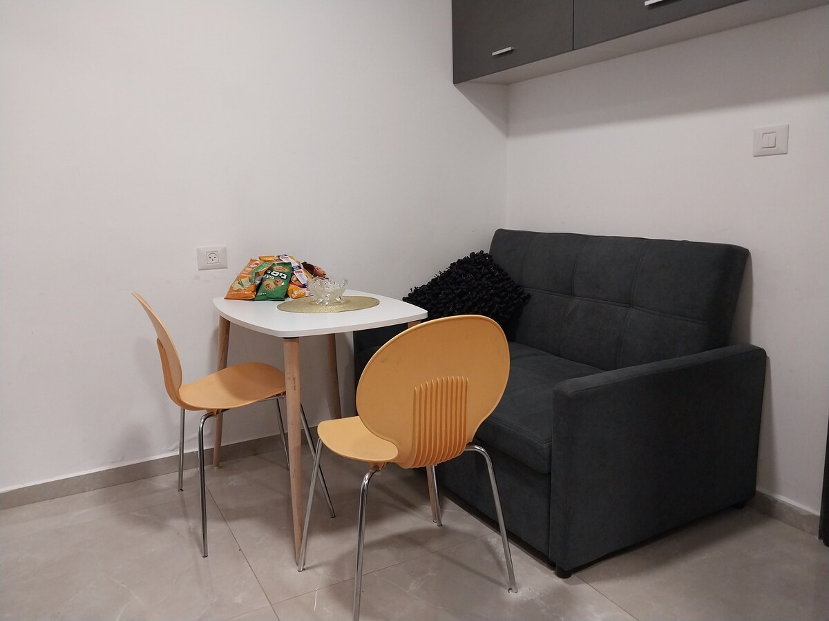 A small dining area is featured, consisting of a round table and two yellow chairs. A cozy gray sofa is present against the wall, complemented by a decorative black cushion. A tray with snacks is placed on the table, enhancing the inviting nature of the space.