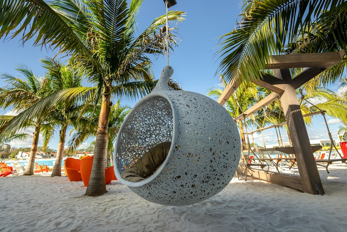 A white hanging chair with intricate patterns is suspended between palm trees on a sandy beach. The chair is cushioned and invites relaxation, with vibrant lounge chairs visible in the background, surrounded by lush tropical greenery.