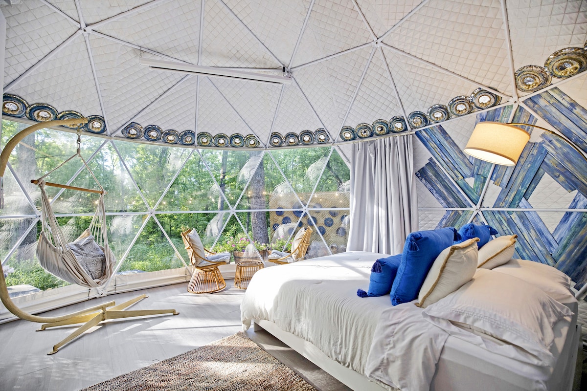 A bright and airy bedroom within the dome features a spacious bed with luxury bedding. A distinctive hanging chair complements the natural light filtering through large geodesic windows. Decorative accents and a neutral rug add to the serene ambiance, surrounded by lush greenery.