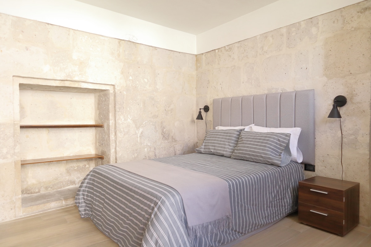 A comfortable sleeping area features a spacious bed with striped bedding. Soft, neutral tones are complemented by the stone wall backdrop. Nightstands with wall-mounted lamps provide gentle lighting, while a simple wooden shelf unit is positioned to one side.