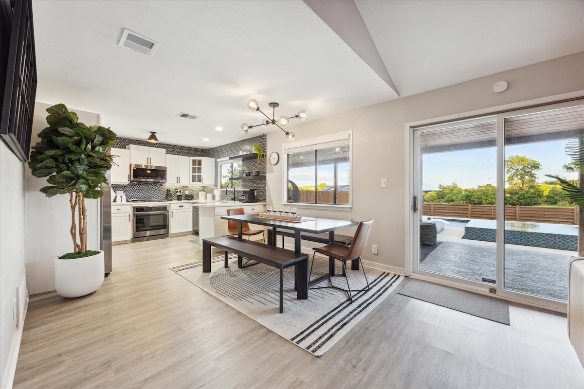 A modern dining area features a long wooden table surrounded by several chairs. Large windows offer views of the pool area, while an open kitchen with contemporary appliances is visible in the background. A decorative plant adds a touch of greenery to the space.