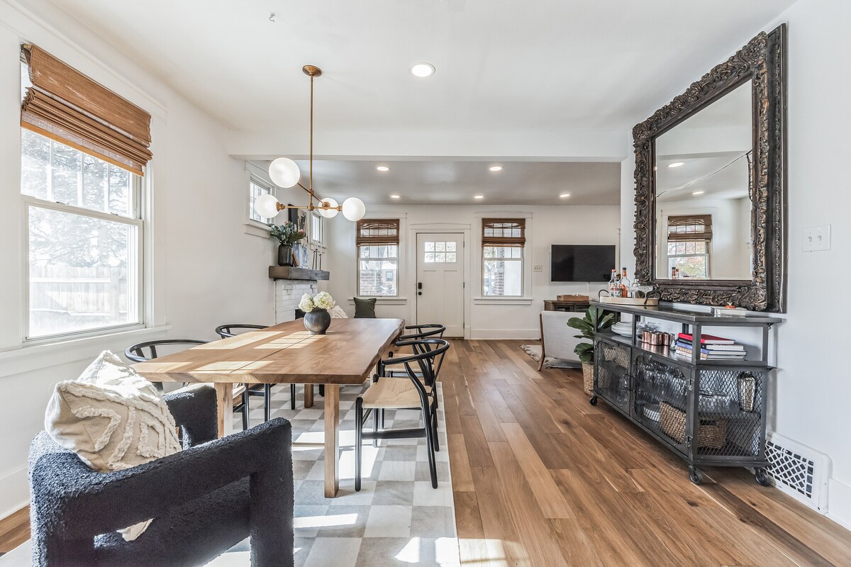 A dining area is shown with a wooden table surrounded by black chairs. Natural light fills the space through several windows with wooden blinds. A large mirror reflects the room, while a cozy seating area is visible in the background.
