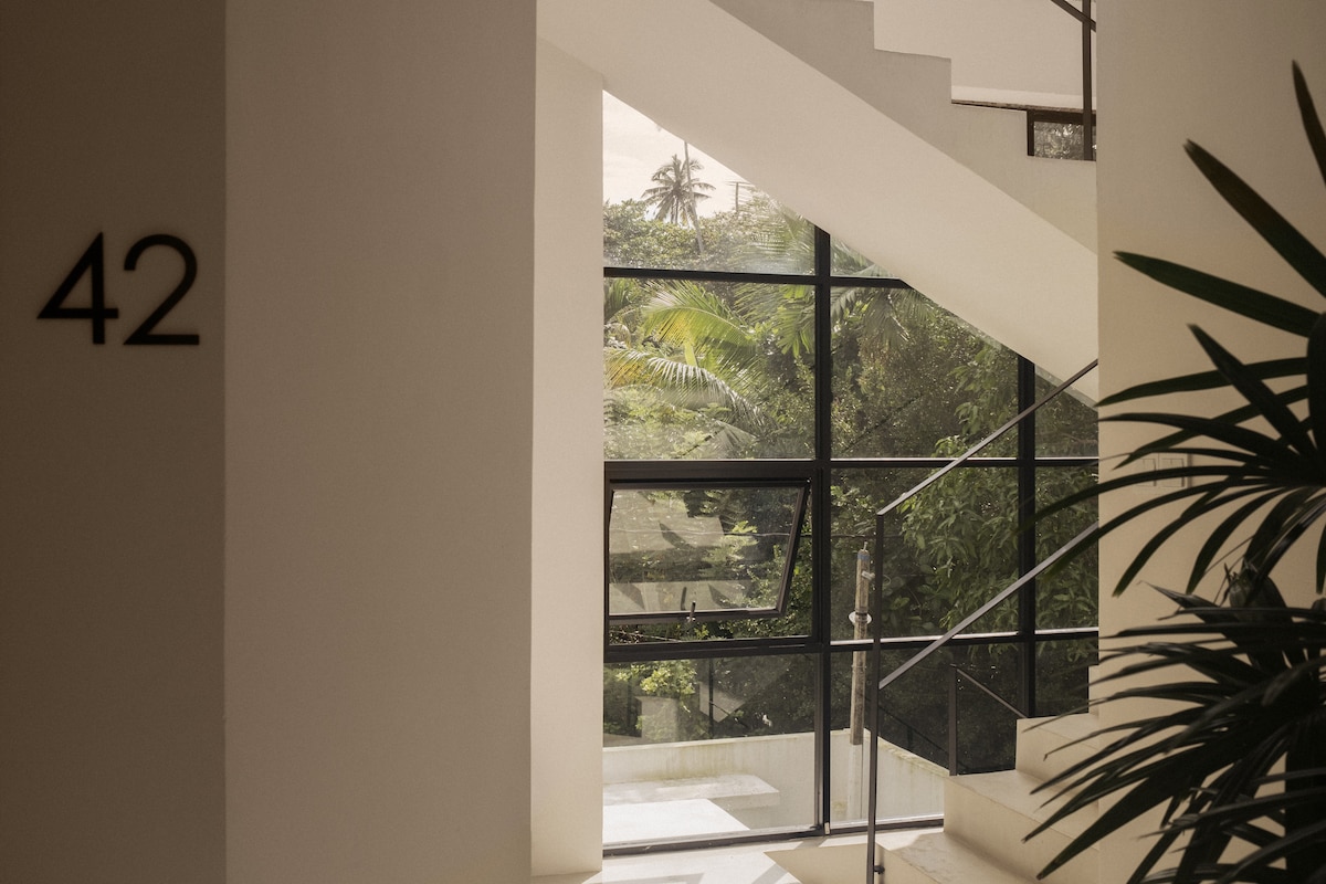 A modern staircase is visible, framed by large glass windows that provide a view of surrounding greenery. The wall features the number '42', and a potted plant adds a touch of nature to the minimalist design.