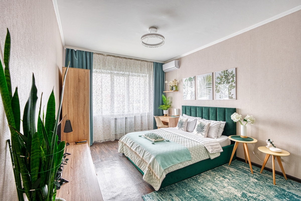 A spacious bedroom features a comfortable green bed with accent pillows. Light curtains allow natural light to filter through the window. A decorative plant adds a touch of greenery, while framed wall art complements the soft color palette. Nightstands with lamps offer convenience.