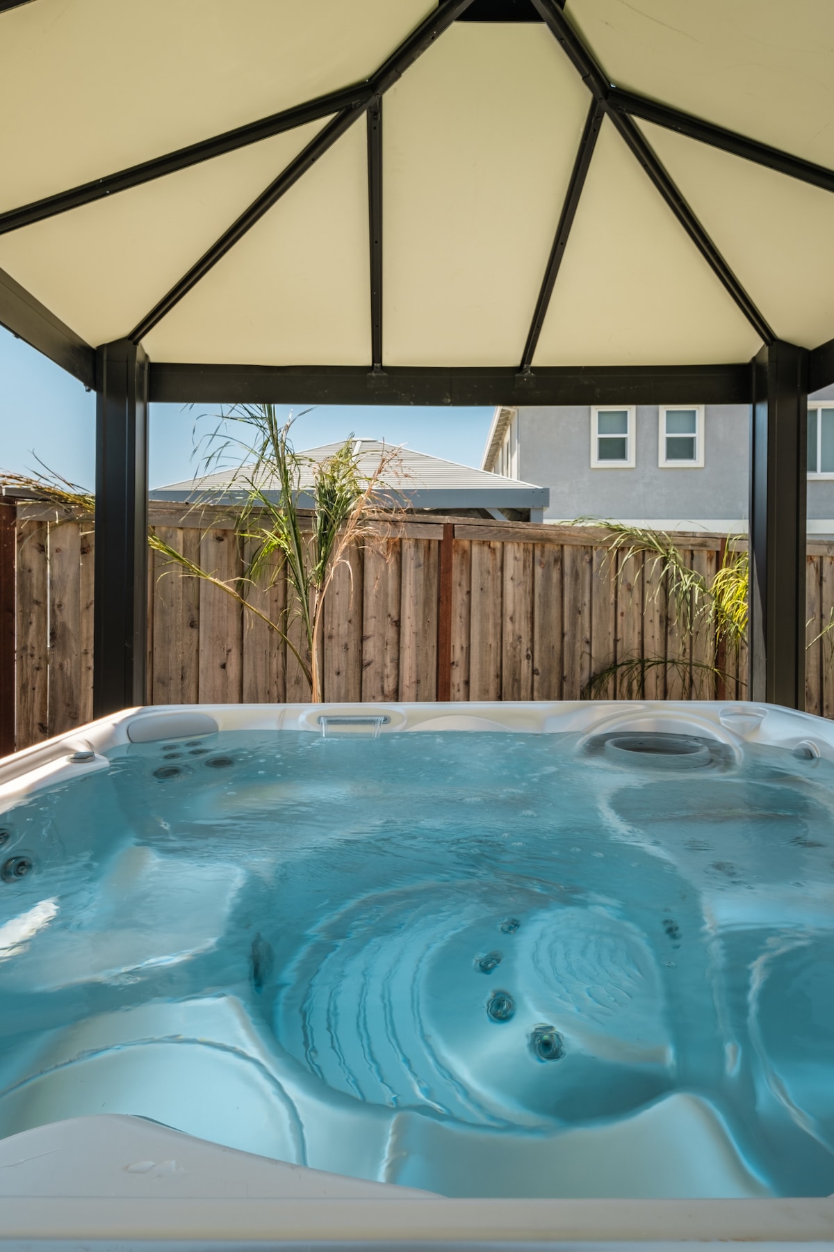 A covered hot tub is situated within a gazebo, featuring clear blue water with visible jets. Surrounding the area are wooden fences and a few plants, providing a sense of privacy and tranquility.